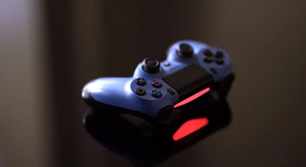 Controle do PS4, DualShock 4, em uma mesa preta, de vidro refletindo o controle azule  preto, ligado com a lightbar do controle exibindo a cor vermelha no LED