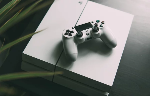 PS4 branco, desligado, com o DusalShock 4 em cima do console em cima de uma mesa de madeira escura.